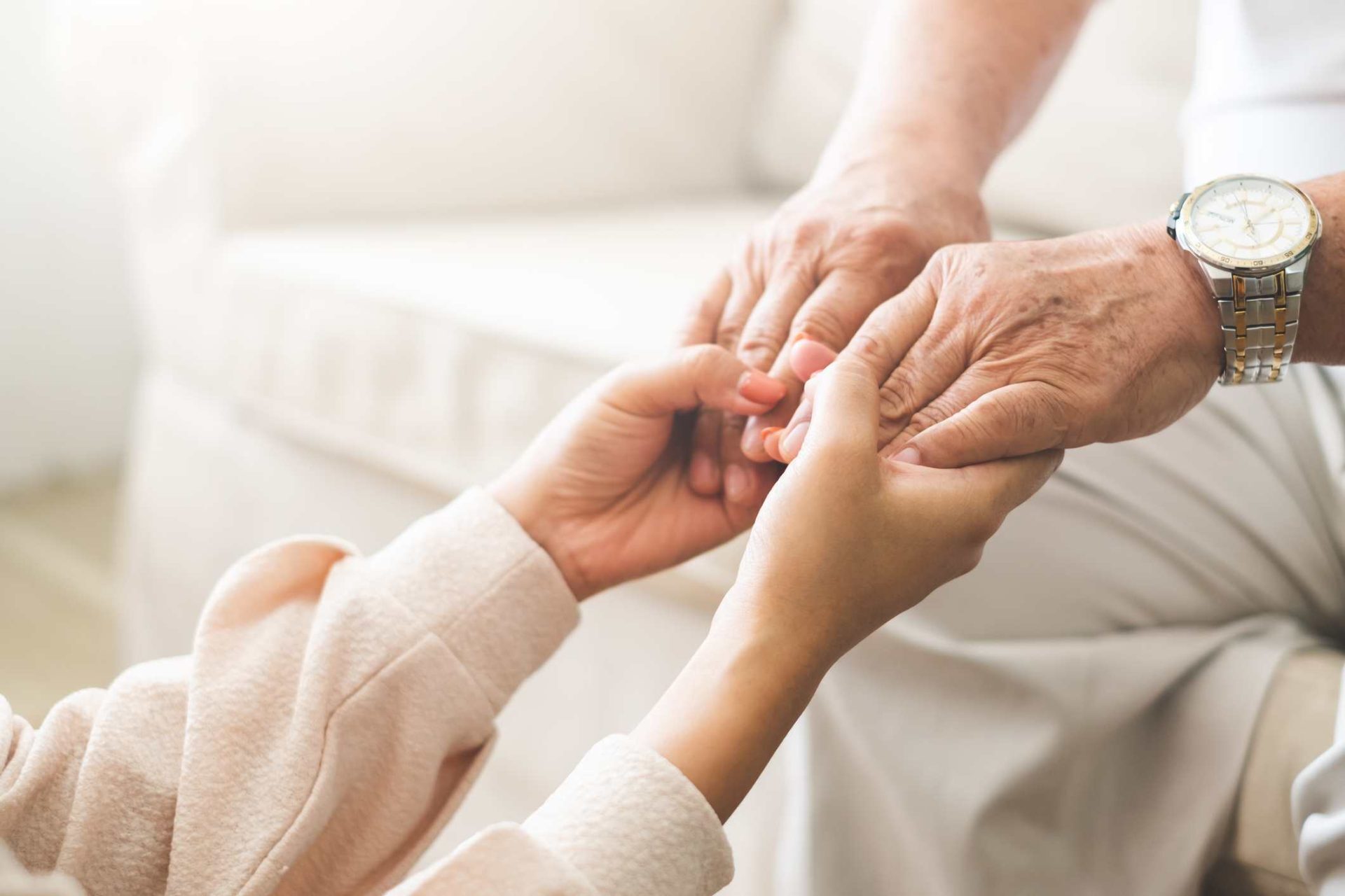 Close-up of a younger person gently holding the hands of an older person wearing a watch, both seated indoors in comfortable environments. Close-up of a younger person gently holding the hands of an older person wearing a watch, both seated indoors in comfortable environments.